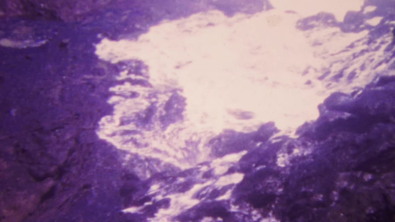 1964 Honolulu home-movie frame showing bright rushing water crossing dark rocky terrain with soft 8mm grain and purple-pink fading.