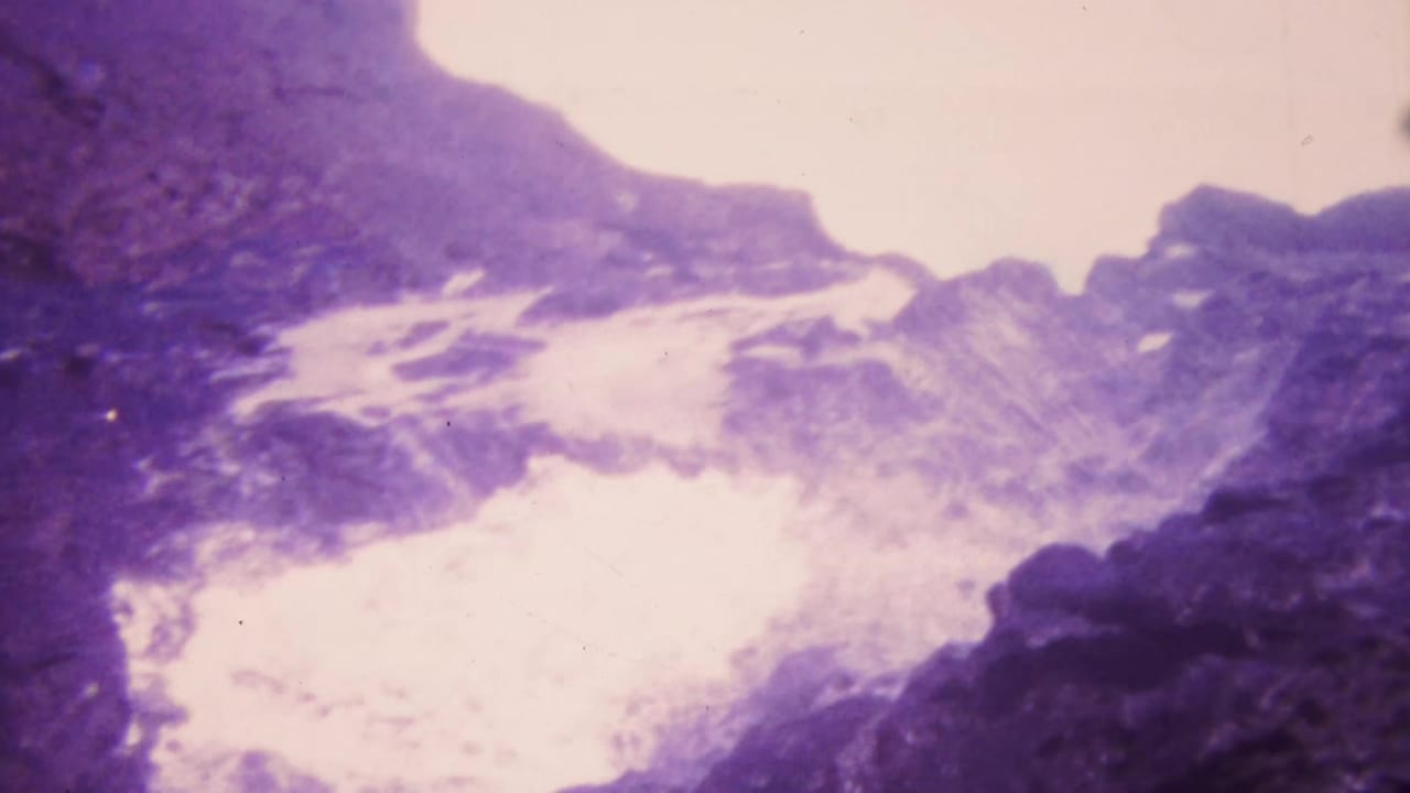 1964 Honolulu footage frame with overexposed rushing water between rugged rocks, carrying the soft focus and magenta shift of aged 8mm film.