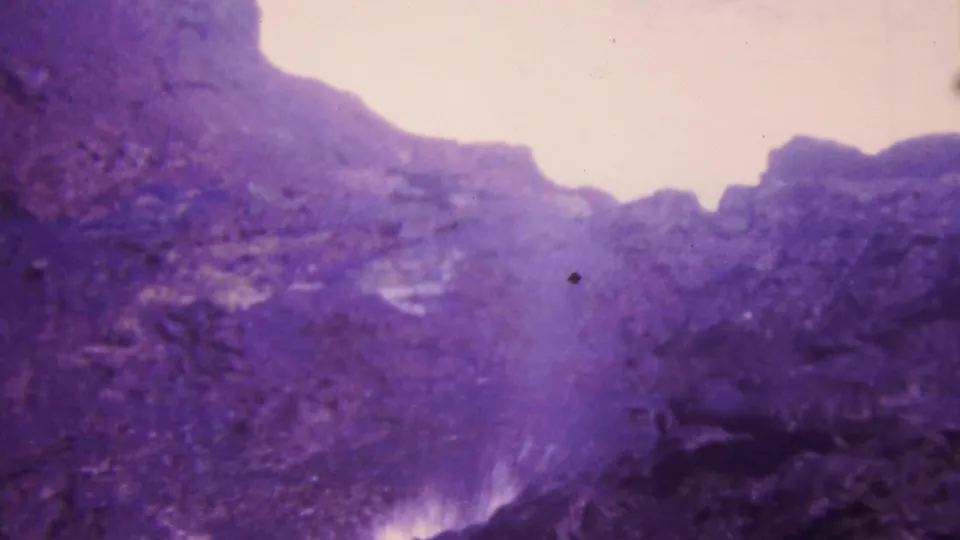 1964 Honolulu home-movie frame looking into a rocky water channel, with pale highlights above, dark wet rock below, and strong purple film fading.
