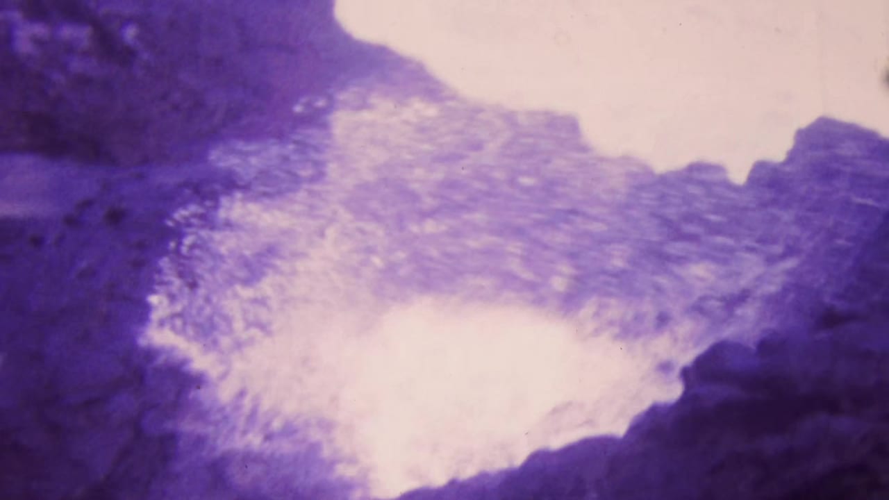 1964 Honolulu 8mm frame with bright surf foam spreading over rough cliff rock, softened by grain and magenta-purple color shift.