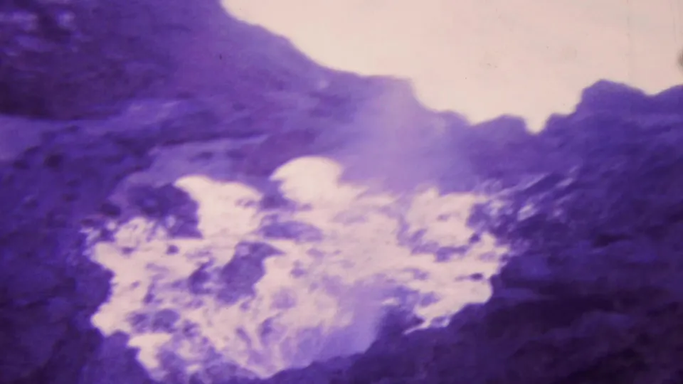 1964 Honolulu 8mm frame showing churning white water below dark shoreline rocks, with aged-film softness and purple-blue coloration.