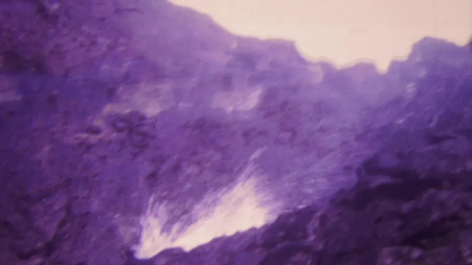 1964 Honolulu 8mm frame showing surf spray and white foam hitting a rocky coastal edge, with soft focus and pronounced purple film aging.