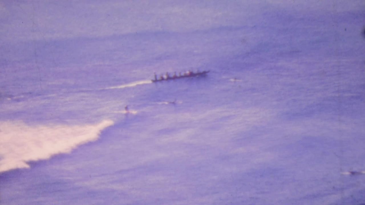 A distant long outrigger canoe with multiple paddlers moves across purple-hued Honolulu ocean water in 1964 home-movie footage, with white wake and small surf visible nearby.