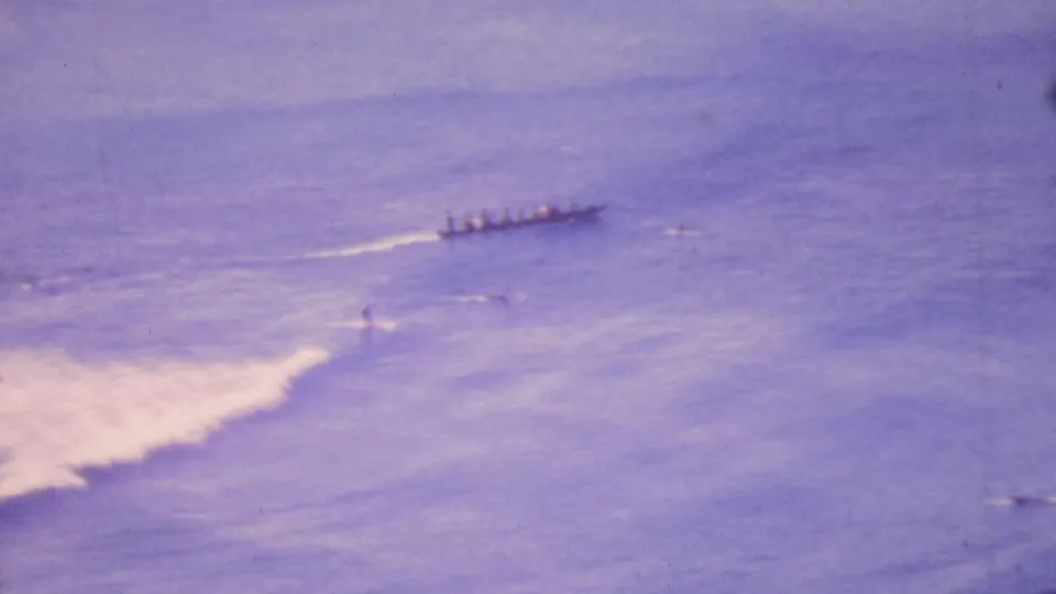 A distant long outrigger canoe with multiple paddlers moves across purple-hued Honolulu ocean water in 1964 home-movie footage, with white wake and small surf visible nearby.
