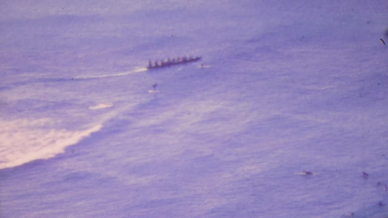 The outrigger canoe continues across the calm purple-blue ocean in Honolulu, with paddlers seated in a line and a pale wake trailing behind the vessel.