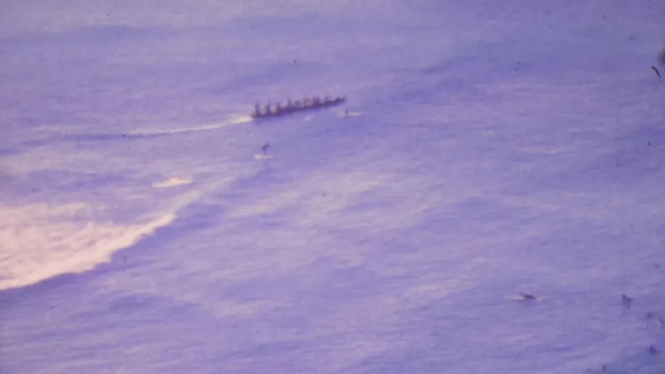 The outrigger canoe continues across the calm purple-blue ocean in Honolulu, with paddlers seated in a line and a pale wake trailing behind the vessel.