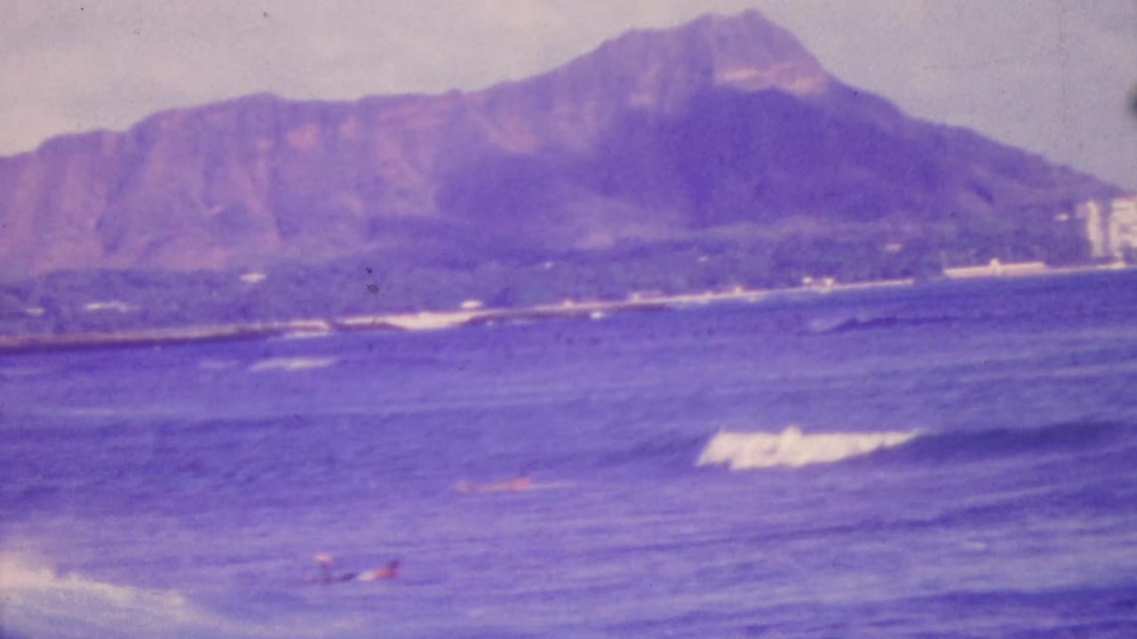 1964 Honolulu home-movie frame showing Diamond Head beyond blue-purple ocean water, with distant swimmers and surf in the foreground.