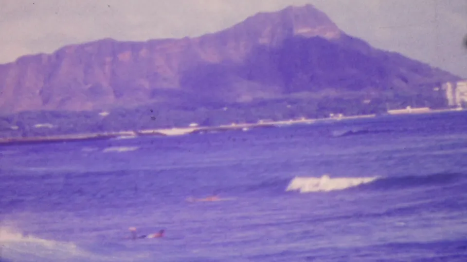 1964 Honolulu home-movie frame showing Diamond Head beyond blue-purple ocean water, with distant swimmers and surf in the foreground.