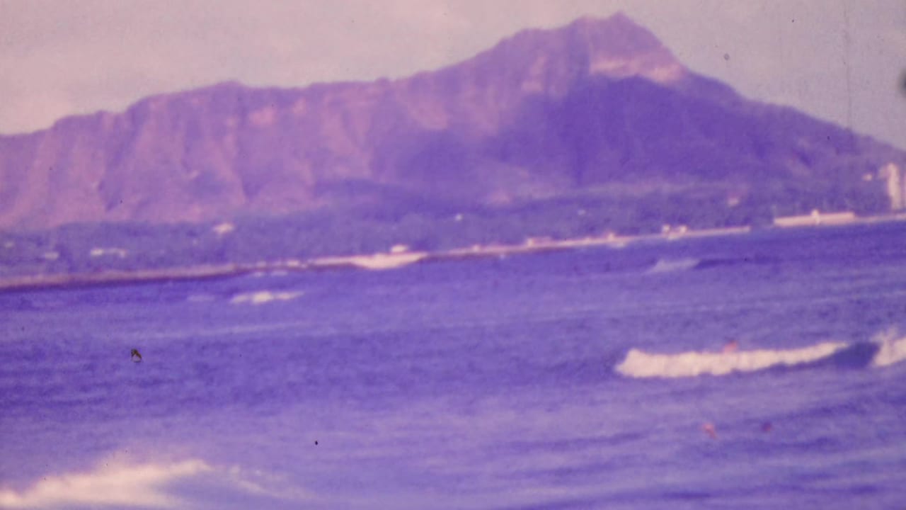 1964 Honolulu 8mm frame with Diamond Head across the bay, purple-shifted ocean water, low surf, and distant beachfront structures.