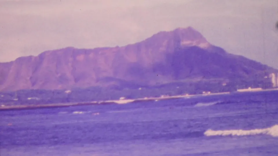 1964 Honolulu home-movie still showing Diamond Head, a distant coastal strip, and rolling blue-purple ocean water with soft 8mm texture.