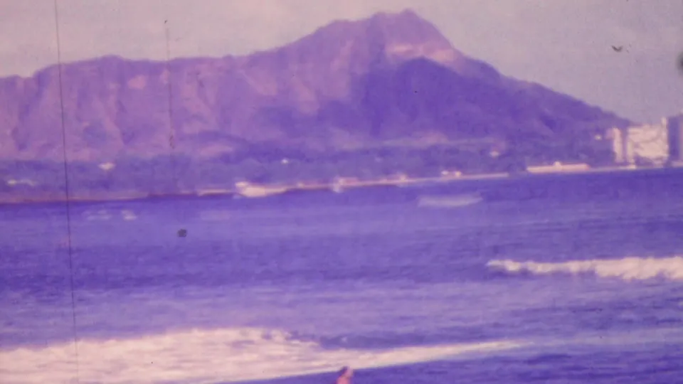 1964 Honolulu 8mm frame showing foreground surf, Diamond Head across the water, and distant shoreline buildings under a pale sky.