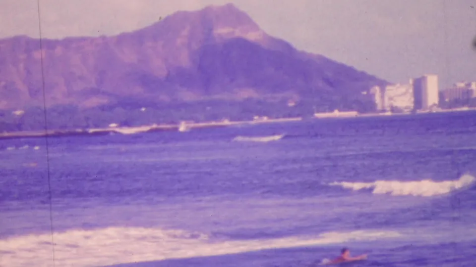 1964 Honolulu home-movie frame showing Diamond Head beyond blue ocean, breaking waves, and distant coastal high-rises with a purple film tint.