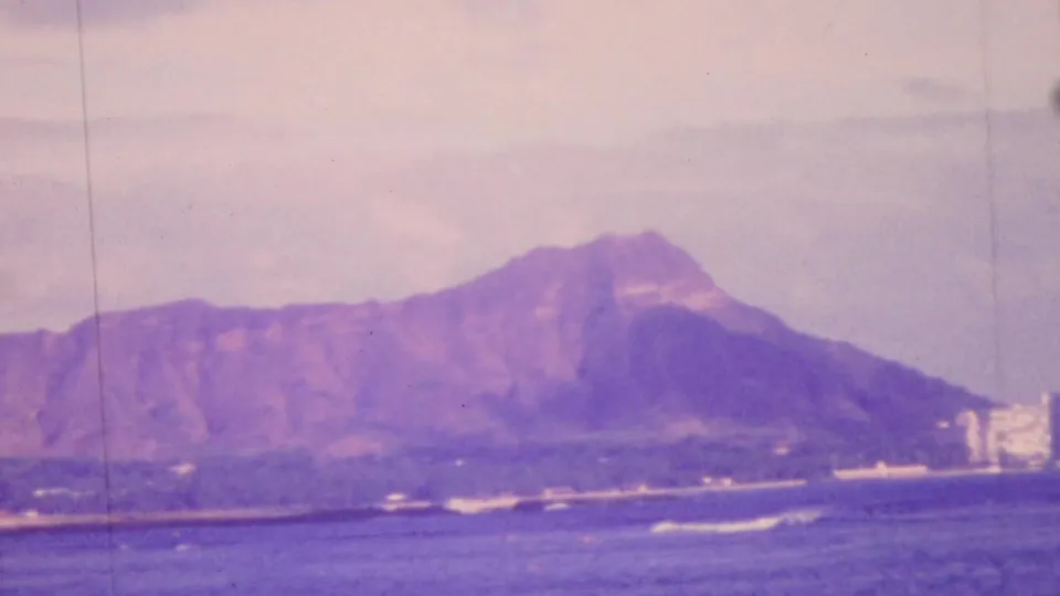 1964 Honolulu home-movie still showing Diamond Head in soft focus with ocean swell, white foam, shoreline structures, and a pronounced purple-blue film cast.