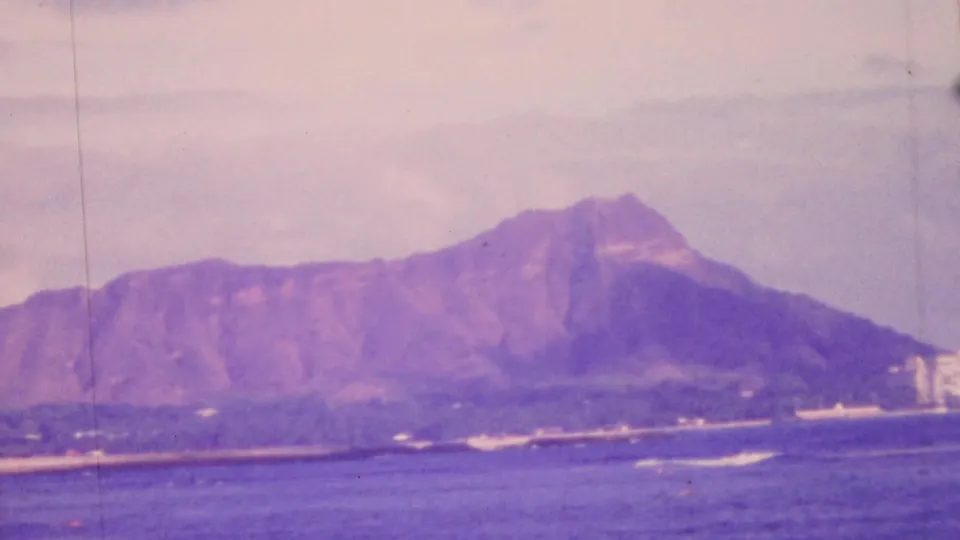 1964 Honolulu frame looking across blue ocean toward Diamond Head, with a low coastal edge, breaking waves, distant buildings, and aged 8mm color shift.