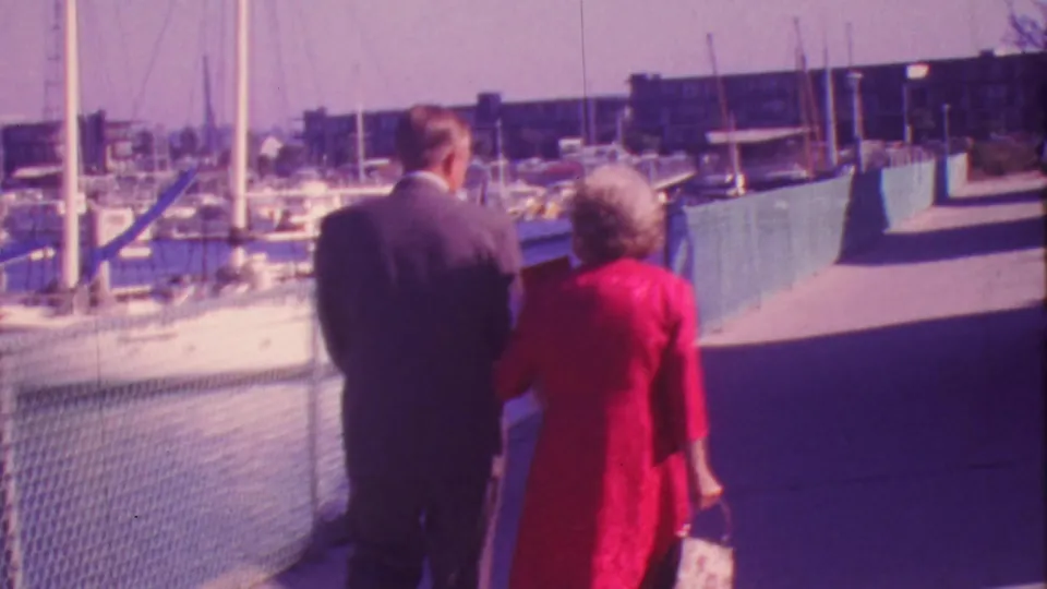 1964 Honolulu home-movie frame showing a man in a dark suit and a woman in a red dress walking away along a marina boardwalk with sailboats nearby.
