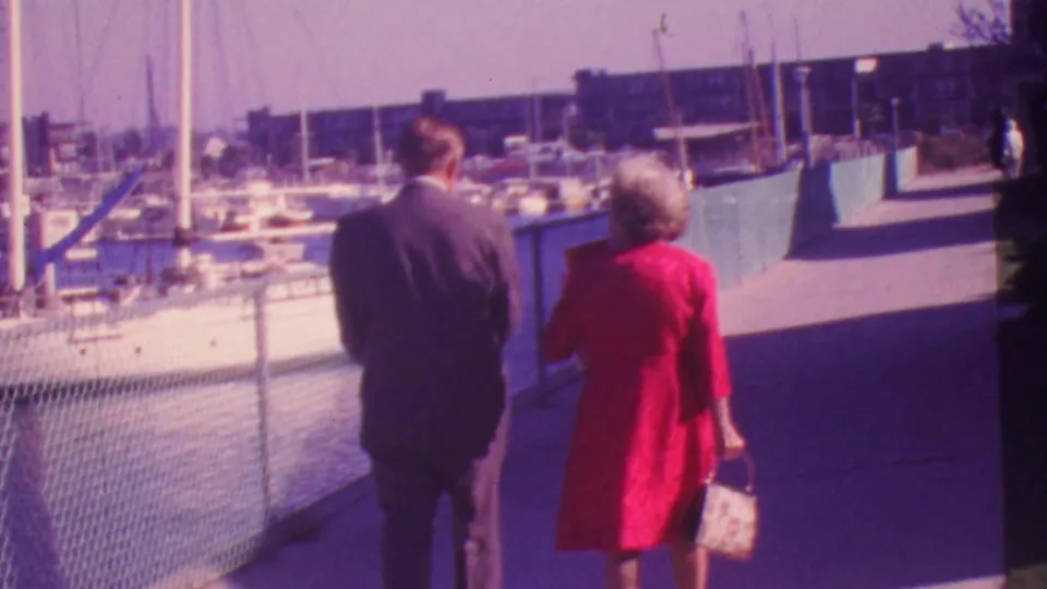 1964 Honolulu marina scene with a couple walking away from the camera beside boats, railings, and waterfront buildings in soft 8mm color.