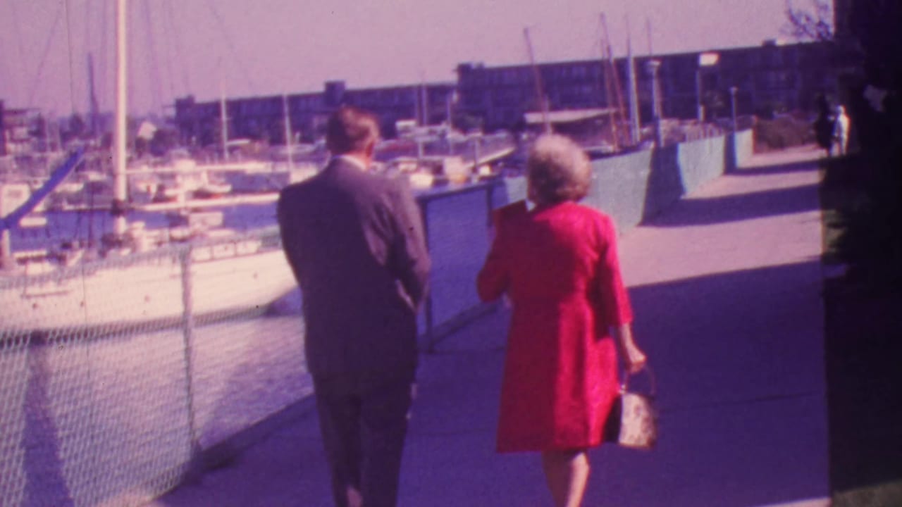 In 1964 Honolulu, a formally dressed couple is seen from behind walking along a sunlit marina walkway beside moored boats.