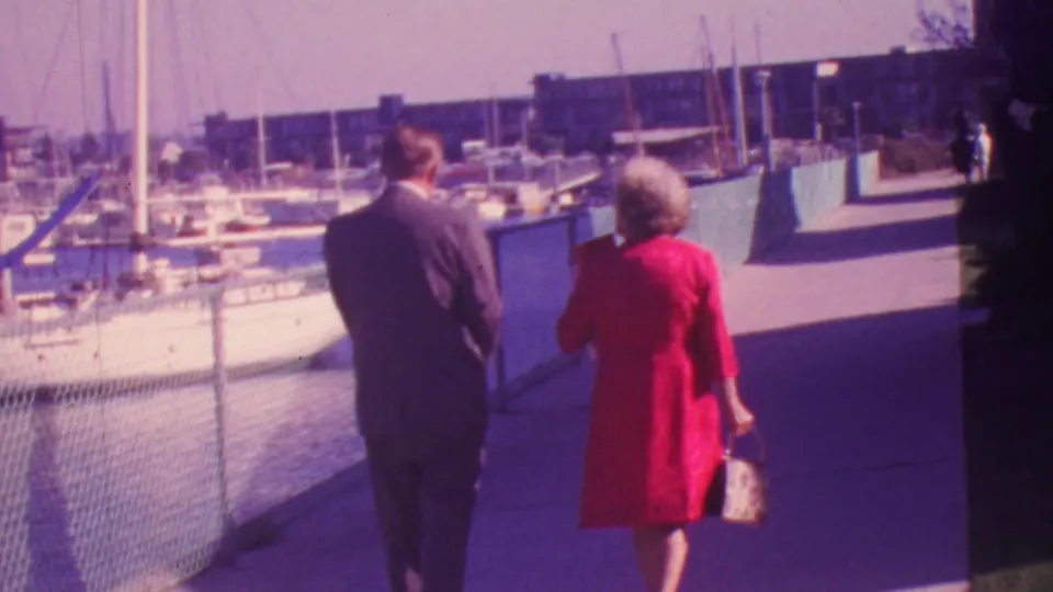 In 1964 Honolulu, a formally dressed couple is seen from behind walking along a sunlit marina walkway beside moored boats.