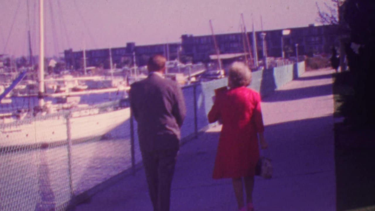 The couple continues along the waterfront walkway with sailboats and buildings beyond the fence.