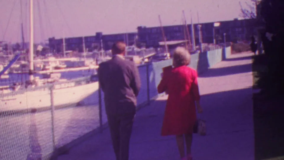 The 1964 Honolulu marina scene continues as the suited man and woman in a red dress walk away from the camera past sailboats and low buildings.