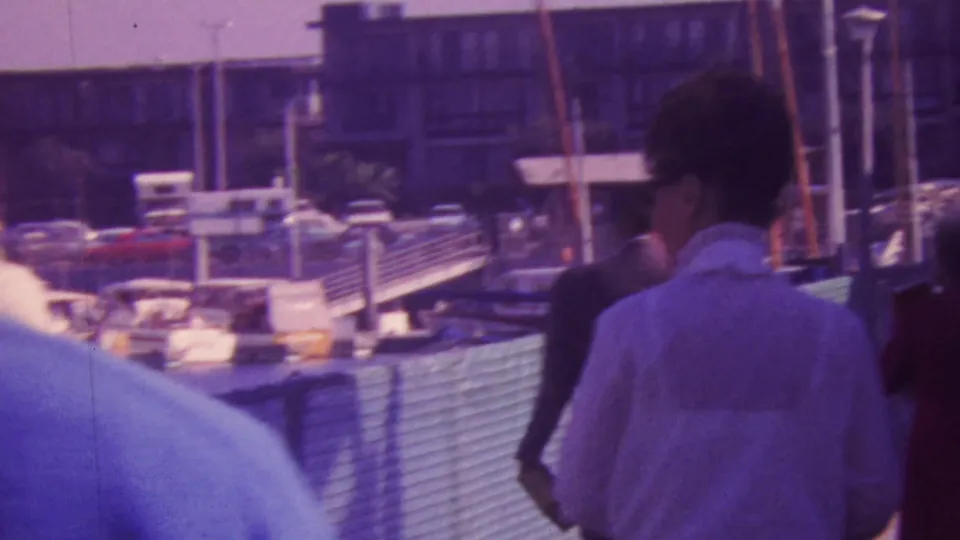 Adults on a Honolulu marina walkway are filmed close to the camera, with moored boats, masts, fencing, and waterfront buildings behind them.