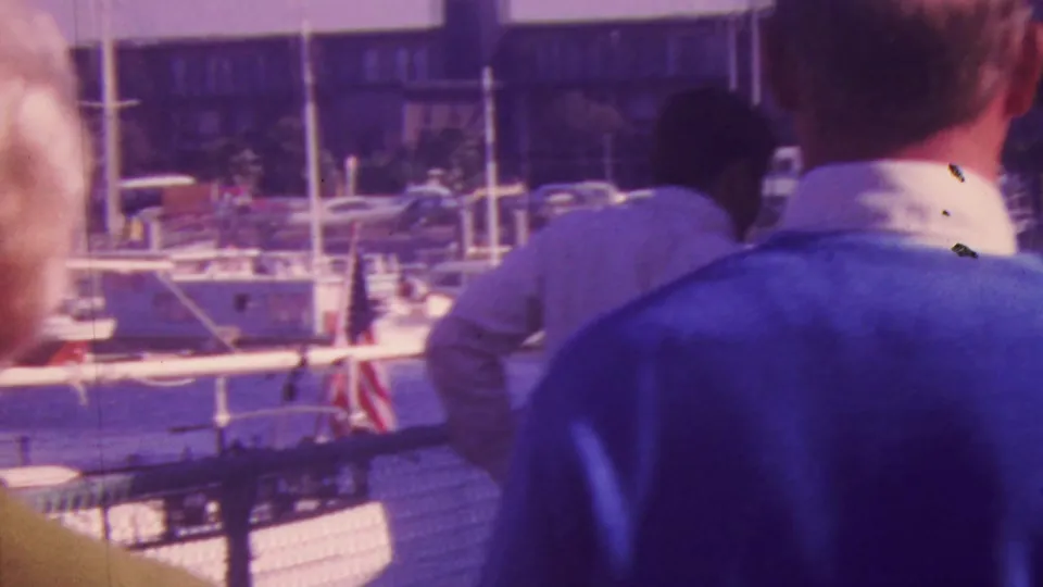 At the Honolulu marina, adults are seen from close range near the fence, including a man in a blue sweater, with sailboats and masts beyond.