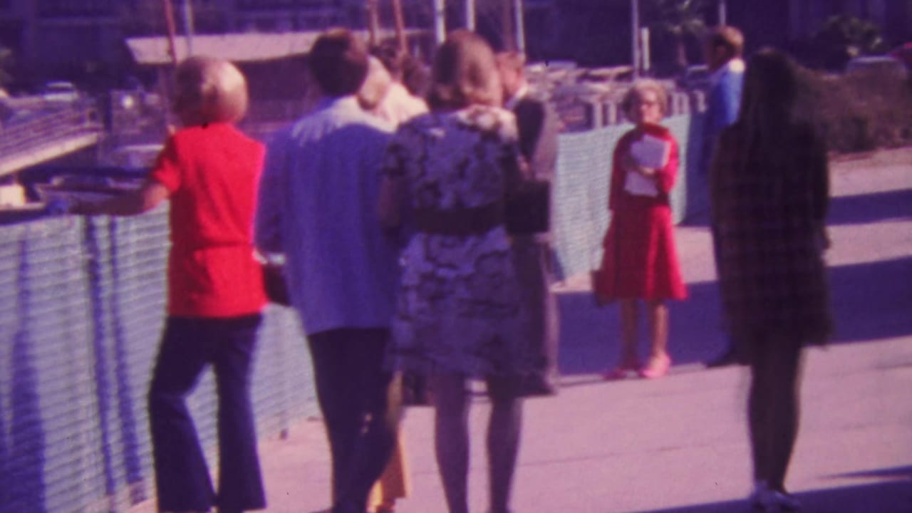 1964 Honolulu home-movie frame showing adults in bright and patterned 1960s clothing walking along a marina-side boardwalk with railings and boats in the background.