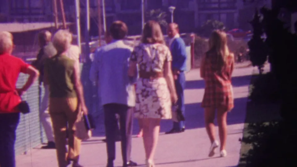 1964 Honolulu boardwalk frame showing adults from behind, including women in floral and plaid dresses and men in light shirts, with marina railings and boats nearby.
