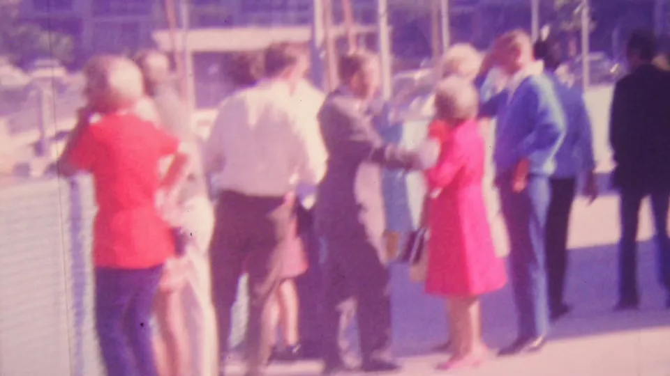Soft 1964 Honolulu home-movie frame with adults meeting or passing on a sunny waterfront boardwalk, including a woman in a red dress and a man in a blue suit.
