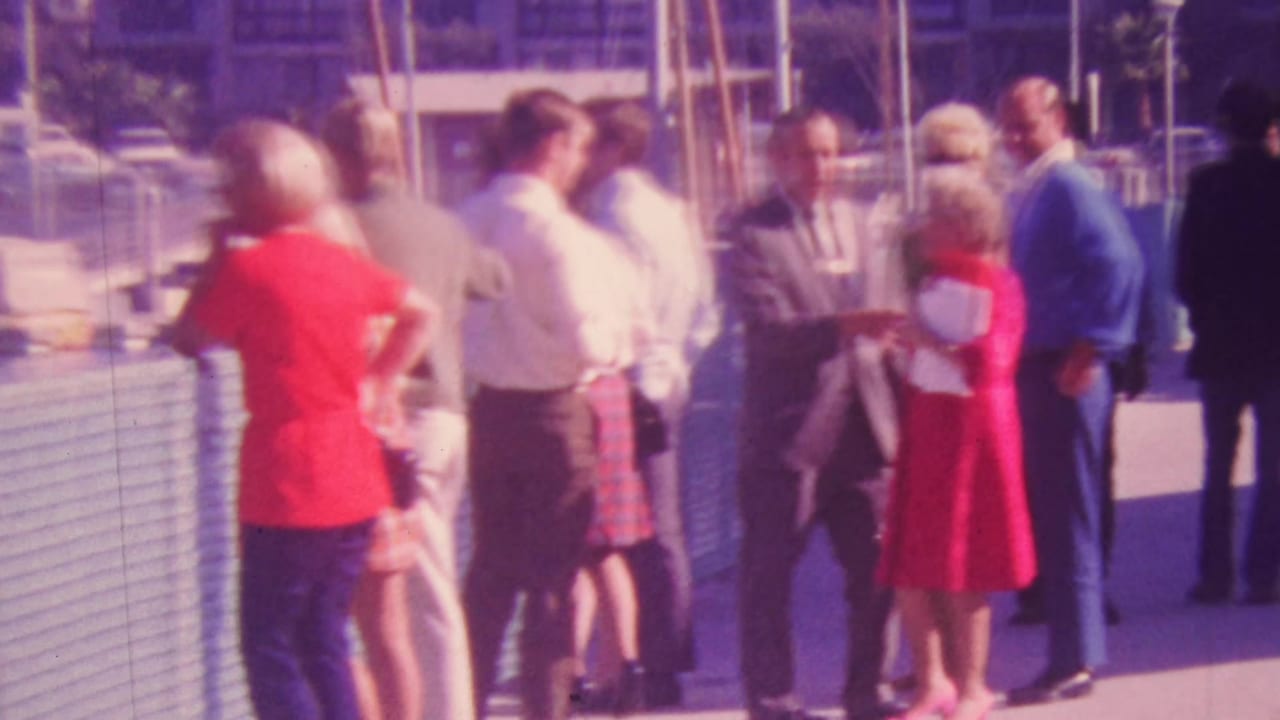 1964 Honolulu home-movie frame showing adults in mid-century clothing gathered near a pier or marina fence, with boats and parked cars in the background.