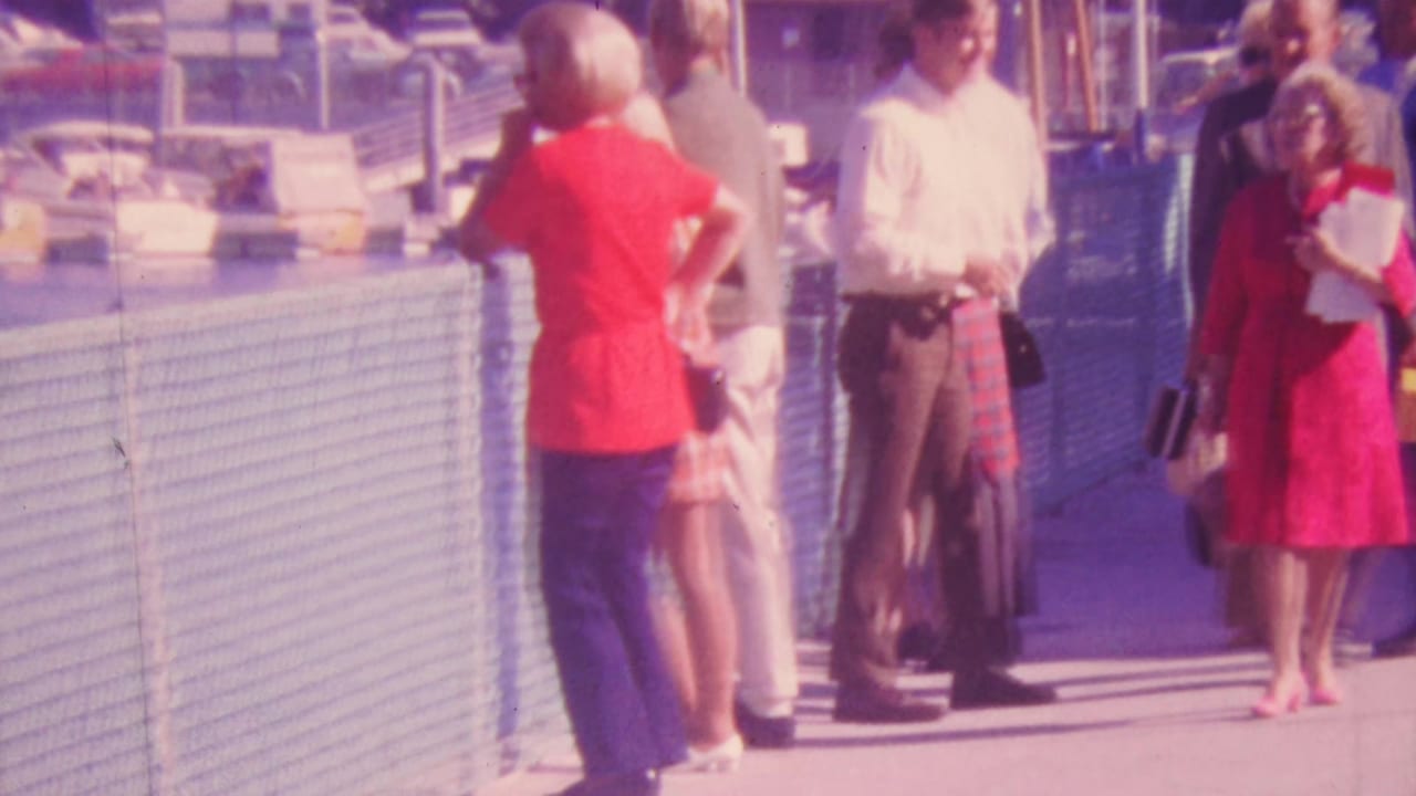 People standing along a waterfront walkway beside a pale fence, with boats visible beyond it.