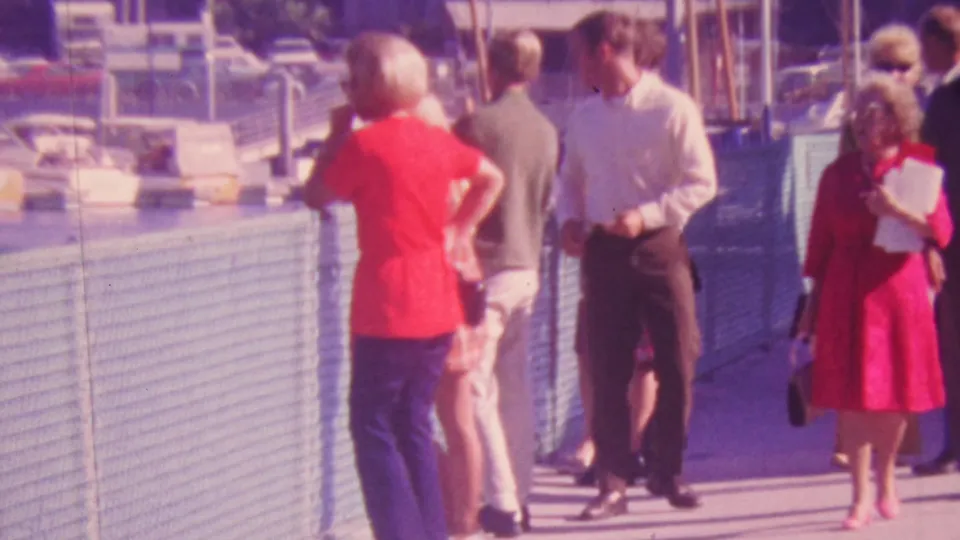 1964 Honolulu pier-side frame showing adults moving through a sunlit public walkway, with a woman in red and men in mid-century casual clothing near the railing.