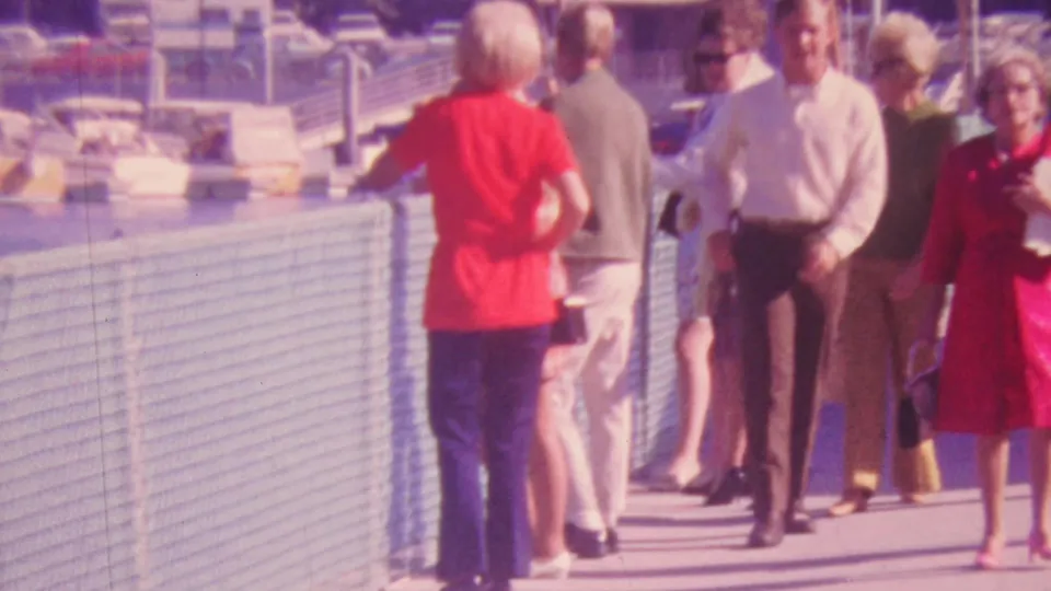 1964 Honolulu home-movie frame showing adults in red clothing, light shirts, and suits near a waterfront fence, with boats and marina details behind them.