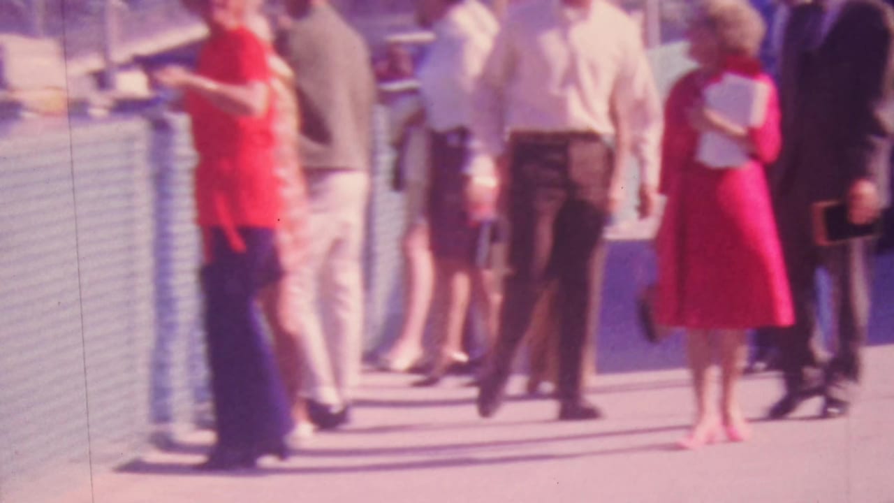 1964 Honolulu home-movie frame showing adults in bright clothing walking along a waterfront pier beside a marina railing and moored boats.
