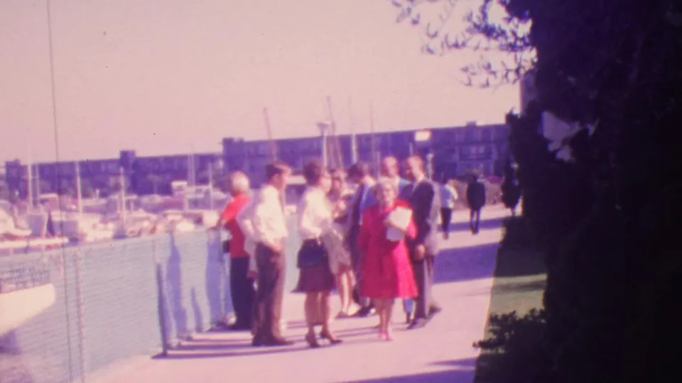 Wide 1964 Honolulu home-movie frame showing a small group pausing on a marina walkway while other pedestrians move through the waterfront scene.