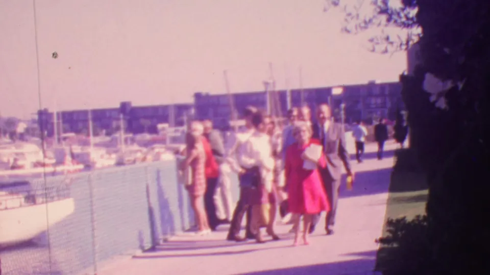 1964 Honolulu home-movie frame showing a mixed-age adult group approaching along a bright marina-side pier, with boats, masts, railing, and tree shadow visible.