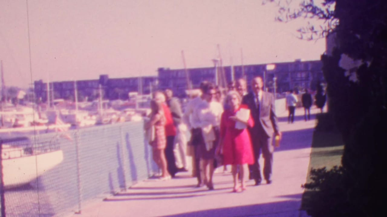 A group of well-dressed adults walks toward the camera on a Honolulu waterfront promenade in 1964, with boats and sailboat masts behind a chain-link fence.