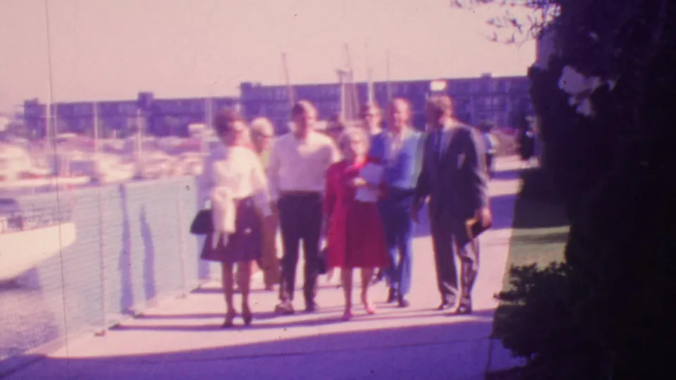 The adult group is nearer the camera on the 1964 Honolulu waterfront, showing dresses, suits, a handbag, the chain-link fence, and sailboat masts in soft 8mm focus.