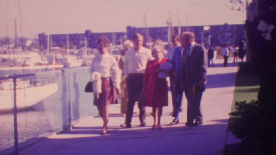 Adults pass through the frame on a Honolulu marina promenade in 1964, with a woman in a red dress, suited men, fence line, water, and sailboat masts visible.