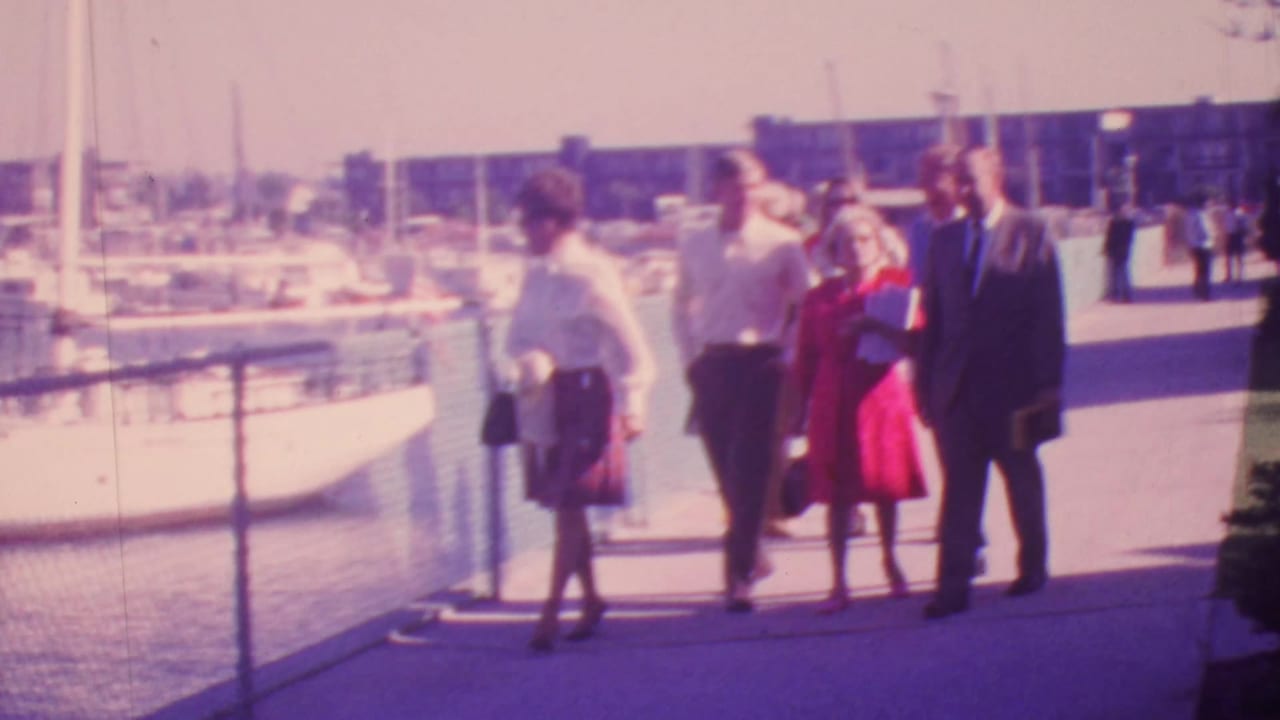 In metadata-supported 1964 Honolulu, several adults walk along a sunny marina walkway with boats, sailboat masts, a chain-link fence, and waterfront buildings behind them.