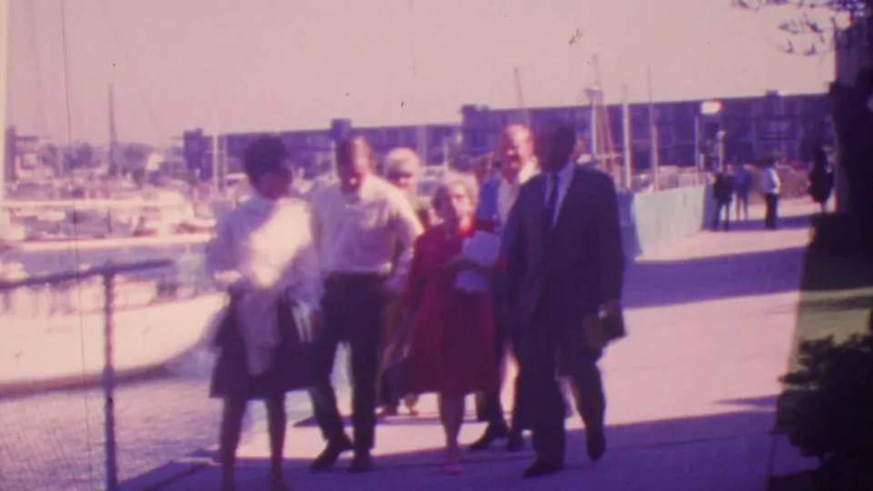 A group of adults, including a woman in a red skirt and a man in a dark suit, walk past moored boats along a Honolulu marina walkway in the 1964 home-movie footage.