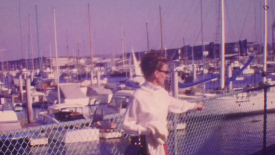 A woman in a white blouse stands or pauses by a chain-link fence at a Honolulu marina, gesturing toward rows of boats and tall sailboat masts visible beyond her.