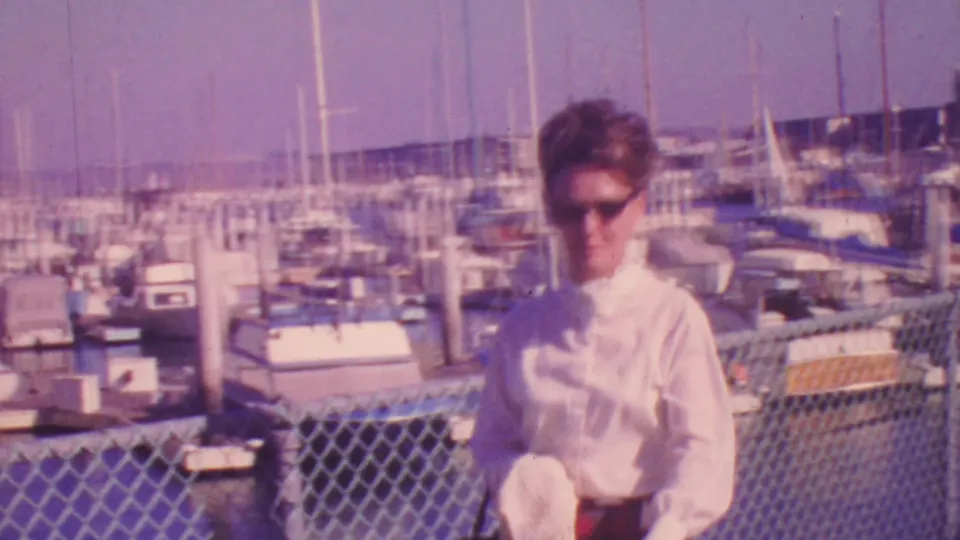 A closer 1964 Honolulu home-movie frame shows a woman in a white blouse and sunglasses near the marina fence, with boats, masts, and dock structures behind her.