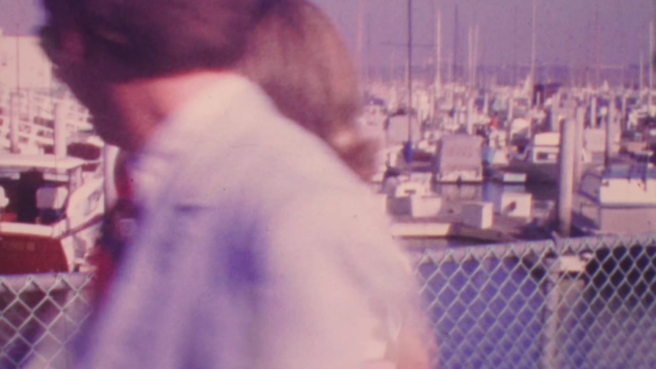 In 1964 Honolulu, a man in a light suit and red tie walks beside a blonde woman along a marina pier with sailboats and masts behind them.