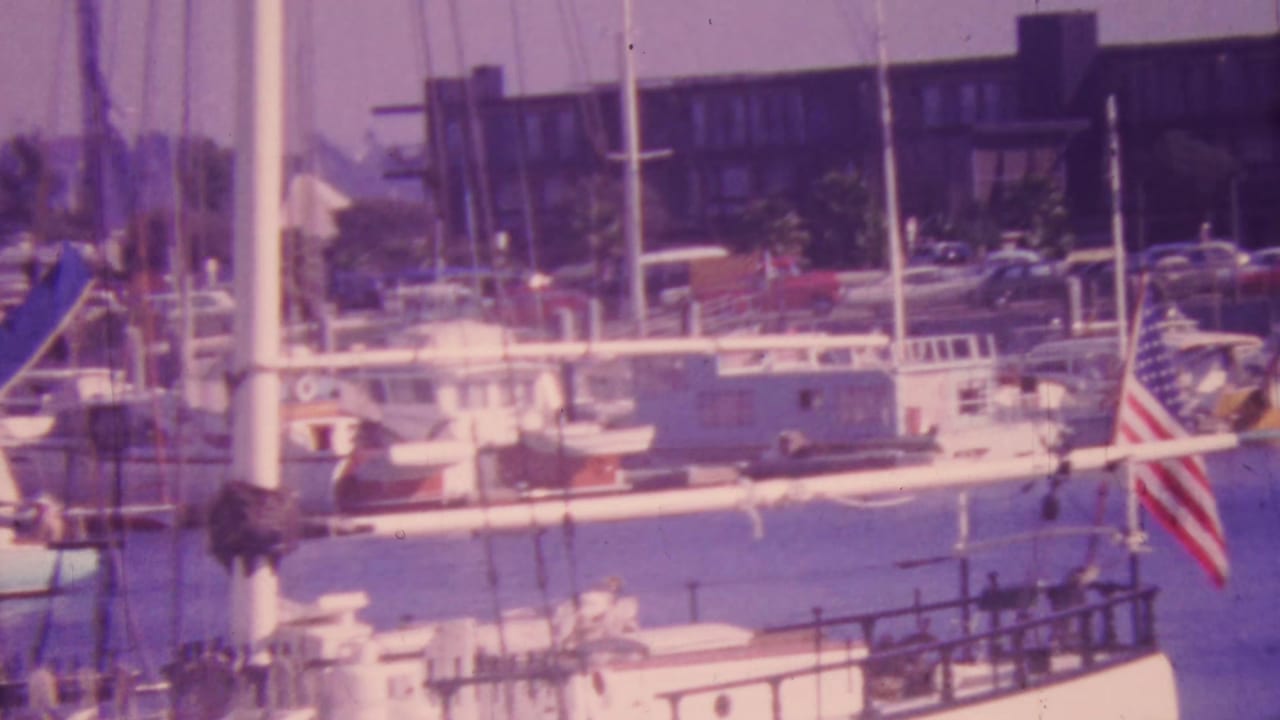Honolulu marina view with docked sailboats, yacht masts, calm harbor water, a waterfront building, parked cars, palm trees, and an American flag.