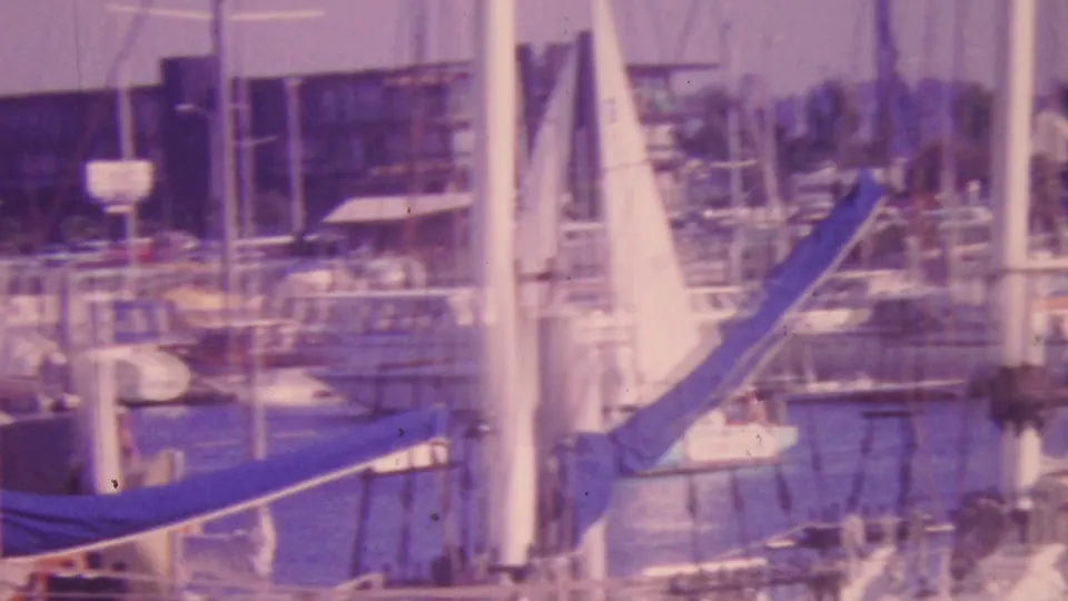 Honolulu marina boats appear through sailboat masts and rigging, with a blue canvas-covered boom, docked vessels, calm water, and a waterfront building in the background.