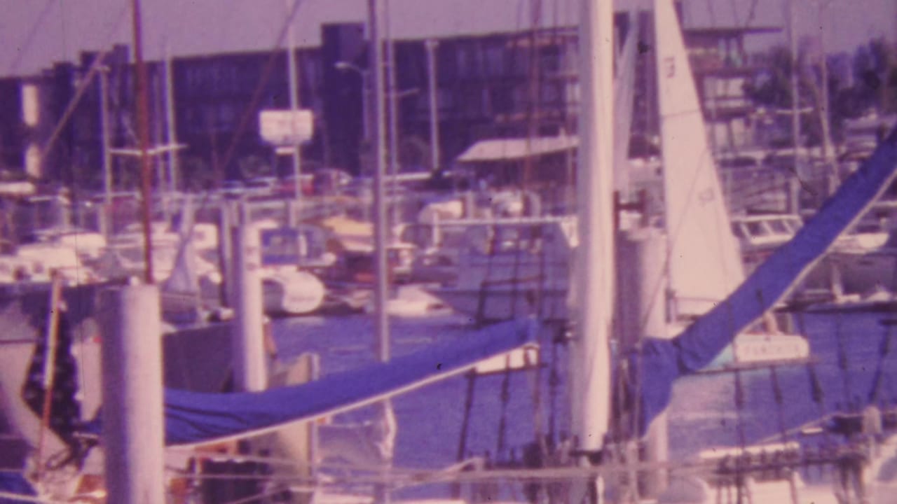 1964 Honolulu marina view with numerous docked sailboats, vertical masts, white pilings, and waterfront buildings in soft faded 8mm color.