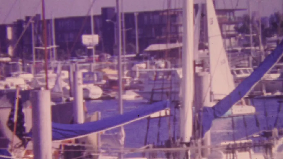 1964 Honolulu marina view with numerous docked sailboats, vertical masts, white pilings, and waterfront buildings in soft faded 8mm color.