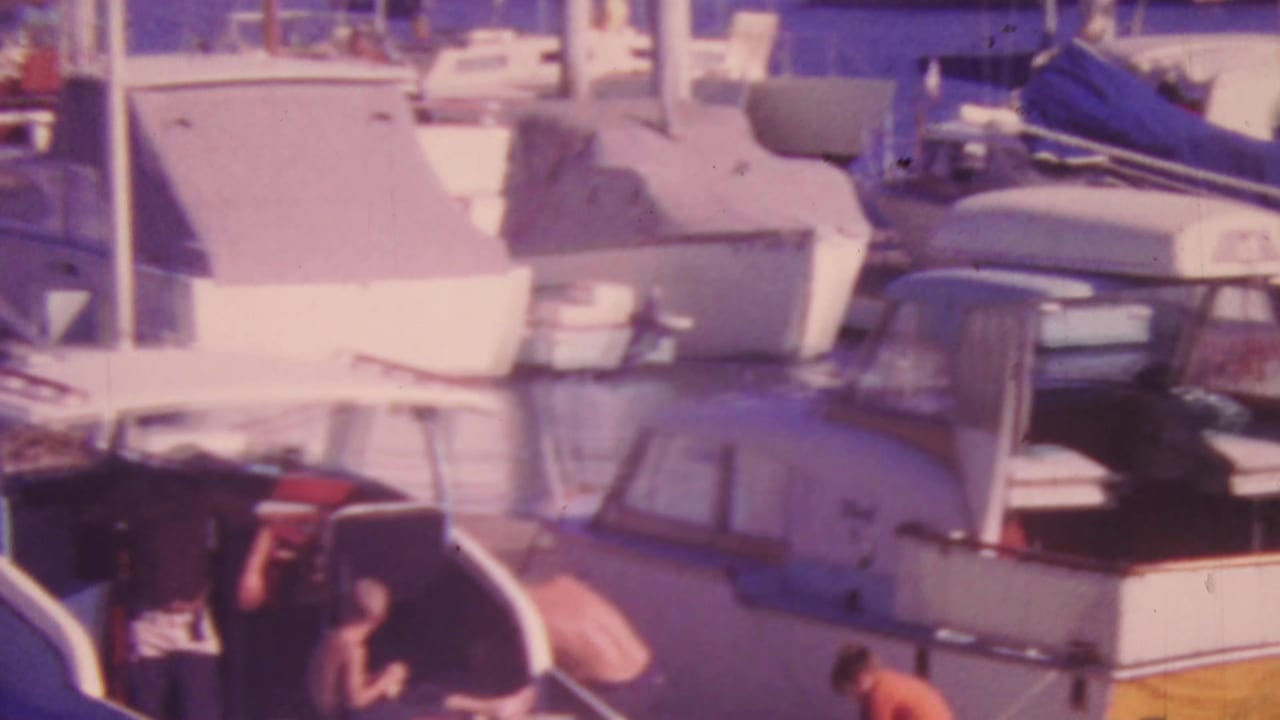 1964 Honolulu marina view with cabin cruisers, sailboats, masts, canvas covers, reflective water, and small indistinct figures near the boats.