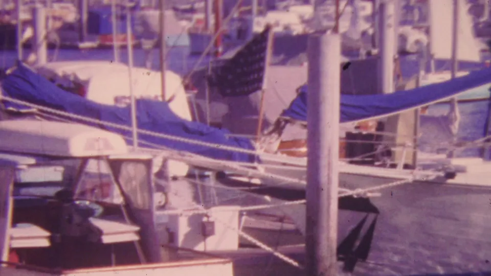 1964 Honolulu marina view with a tall mast in front, sailboat rigging, blue-covered boom, docked cabin cruiser, and water between the slips.